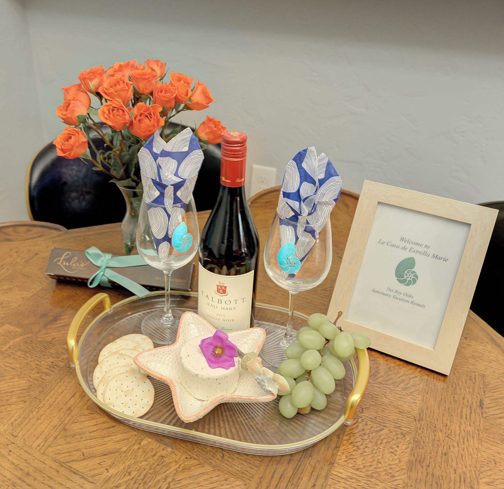A table with a bottle of wine, glasses, a framed welcome sign, a vase of orange roses, and a plate with crackers and grapes.