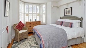 Ein Schlafzimmer mit einem großen Bett, einem Korbstuhl, einem Schminktisch und einem Fenster mit Fensterläden.