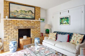 A living room with a brick fireplace, a sofa with patterned cushions, and a painting above the fireplace. - Lovely 2 bed Flat With Garden in Queen s Park (London)