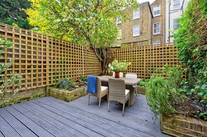 A wooden deck with a table and chairs, surrounded by a wooden fence and greenery. - Lovely 2 bed Flat With Garden in Queen s Park (London)