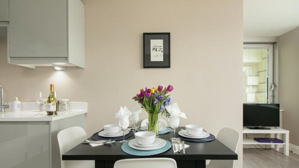 Un comedor con una mesa dispuesta para cuatro personas, un jarrón con flores y un cuadro en la pared.