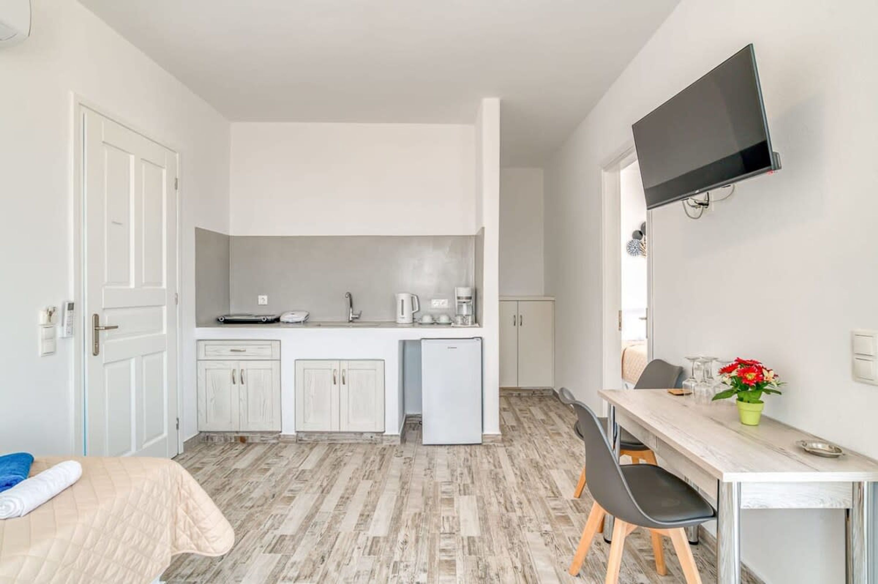 Una cocina compacta con armarios blancos, un refrigerador pequeño y un televisor de pantalla plana montado en la pared.