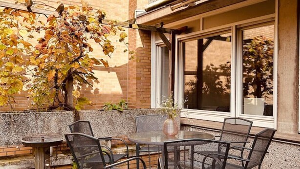 Une terrasse extérieure avec une table ronde et des chaises, un petit vase avec des fleurs et un arbre aux feuilles d’automne.