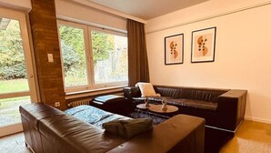 Un salon moderne avec un grand canapé d’angle, une table basse et une cheminée. Deux fenêtres laissent entrer la lumière naturelle et offrent une vue sur l’extérieur.