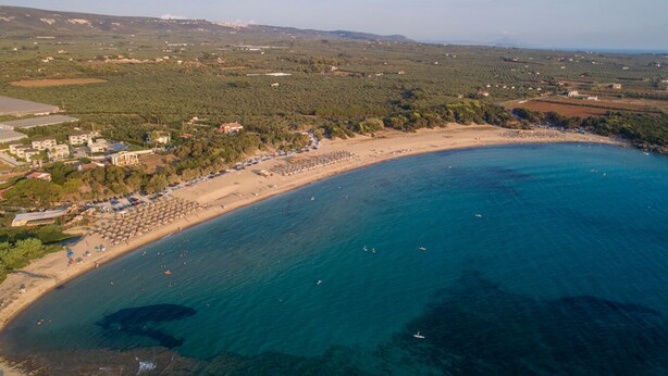 Luchtbeeld van een kustgebied met een strand, helderblauw water en omliggende begroeiing.