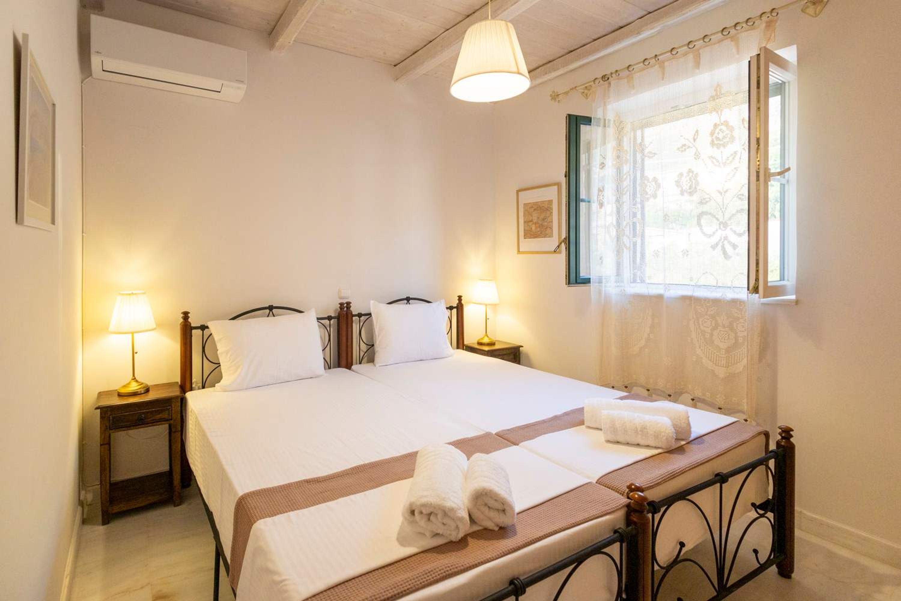 A neatly made bed with white linens and two rolled towels on the bed. There are two bedside tables with lamps, a window with floral curtains, and a picture on the wall.