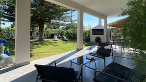 A covered outdoor area with a glass table and chairs, a barbecue grill, and a view of a backyard with a tree and a swing set. - Kalamata Garden Haven - Escape Amidst Greenery (Kalamata)