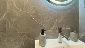 A bathroom with a marble wall, a round mirror with a light, a modern faucet, and a soap dispenser. - Kalamata Garden Haven - Escape Amidst Greenery (Kalamata)