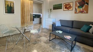 A modern living room with a glass dining table, metal chairs, a grey sofa, and a coffee table. There is a kitchen in the background with white cabinets and a wooden partition. - Kalamata Garden Haven - Escape Amidst Greenery (Kalamata)