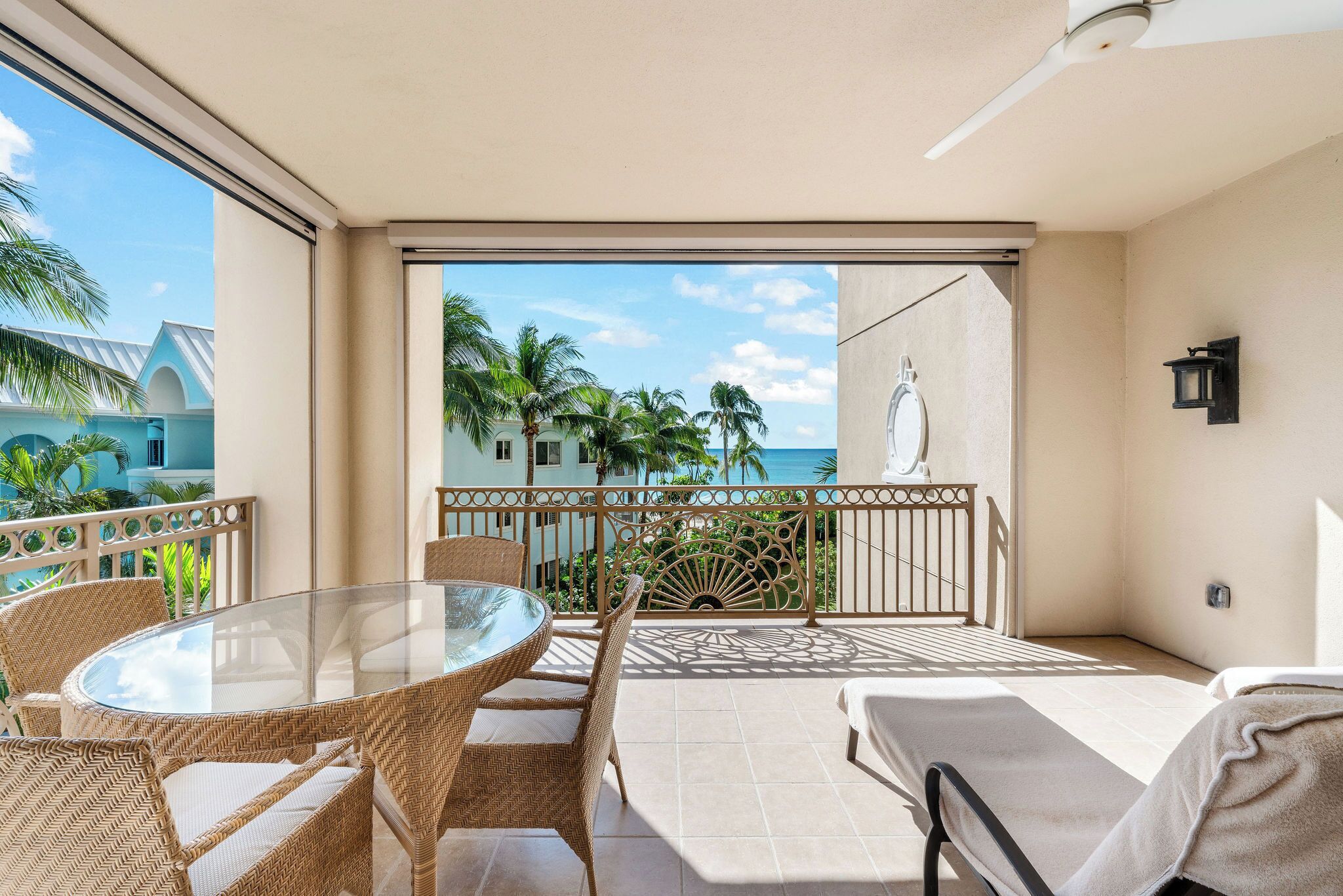 A balcony with a glass table and wicker chairs, overlooking a beach with palm trees and a clear sky.