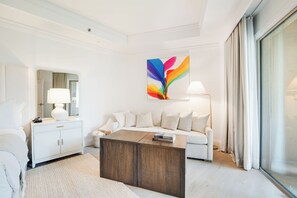 Una habitación de hotel moderna con un sofá blanco, una mesa de centro de madera, un armario blanco, un espejo grande y un cuadro abstracto colorido en la pared.
