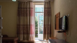 Quarto com televisão sobre suporte, um sofá, janela com cortinas e porta que dá para o exterior.