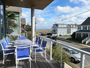 Une terrasse en bois avec une table et des chaises bleues, donnant sur une rue bordée de maisons et de voitures garées.