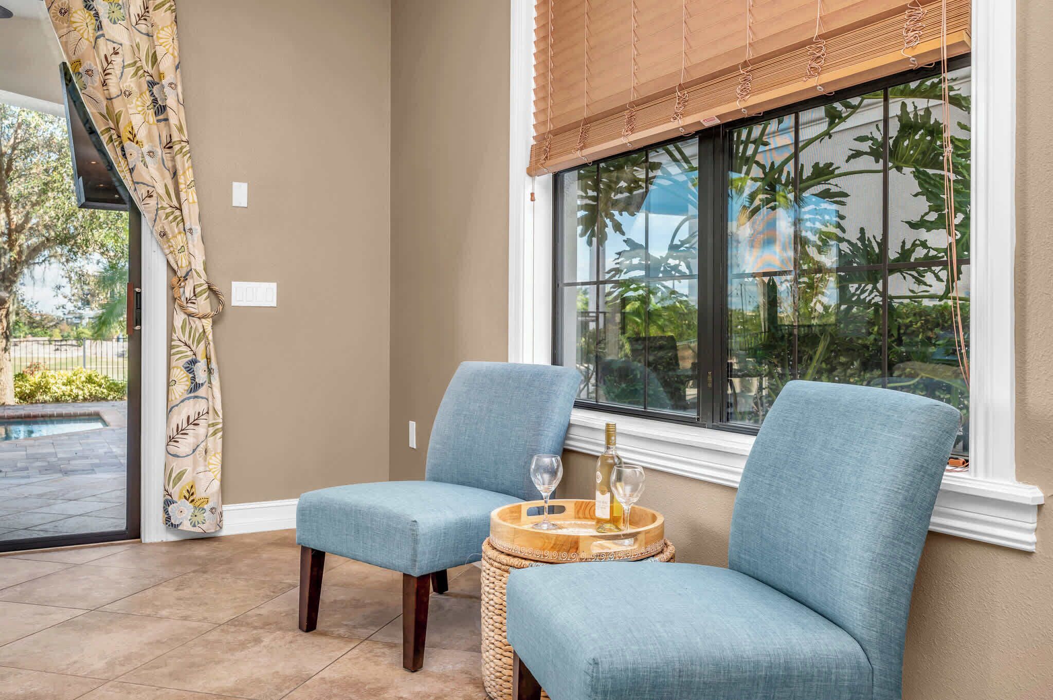 A room with two blue upholstered chairs, a small round table with a bottle and glasses, and a large window with a view of greenery.