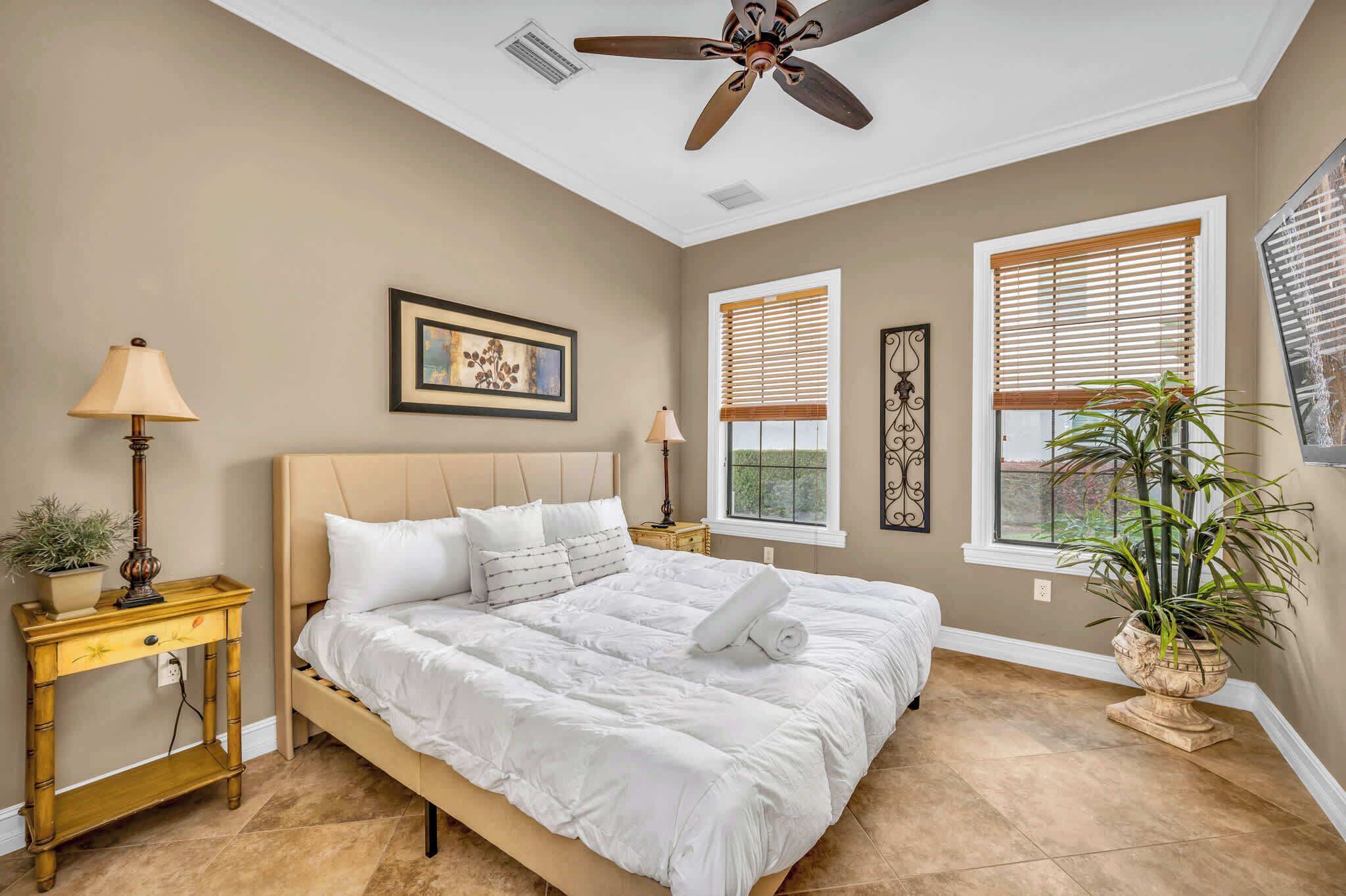 Quarto com cama, duas mesas de cabeceira, ventilador de teto e uma janela grande com persianas.