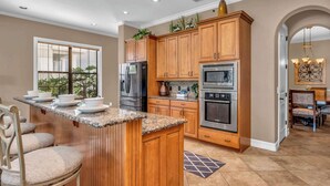 A kitchen with wooden cabinets, granite countertops, a central island, and a dining area with a chandelier. - Bright 5BR retreat w/ pool & fire pit (Kissimmee)