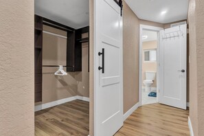 Un couloir doté d’un plancher en bois, une porte blanche avec une poignée noire, et une autre porte blanche donnant sur une salle de bain équipée de toilettes et d’un porte-serviettes.