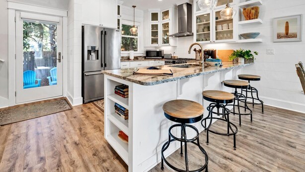 Una cocina moderna con armarios blancos, un refrigerador de acero inoxidable, horno empotrado, una isla grande con taburetes y luminarias colgantes.