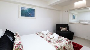 A hotel room with a bed, a chair, a mirror, and a framed picture of a surfer on a wave. - T1-Sunset Apartment (Nazaré)