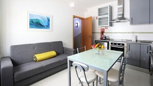 A compact living space with a grey sofa, a glass dining table, and a kitchen area including a stove and cabinets. - T1-Sunset Apartment (Nazaré)