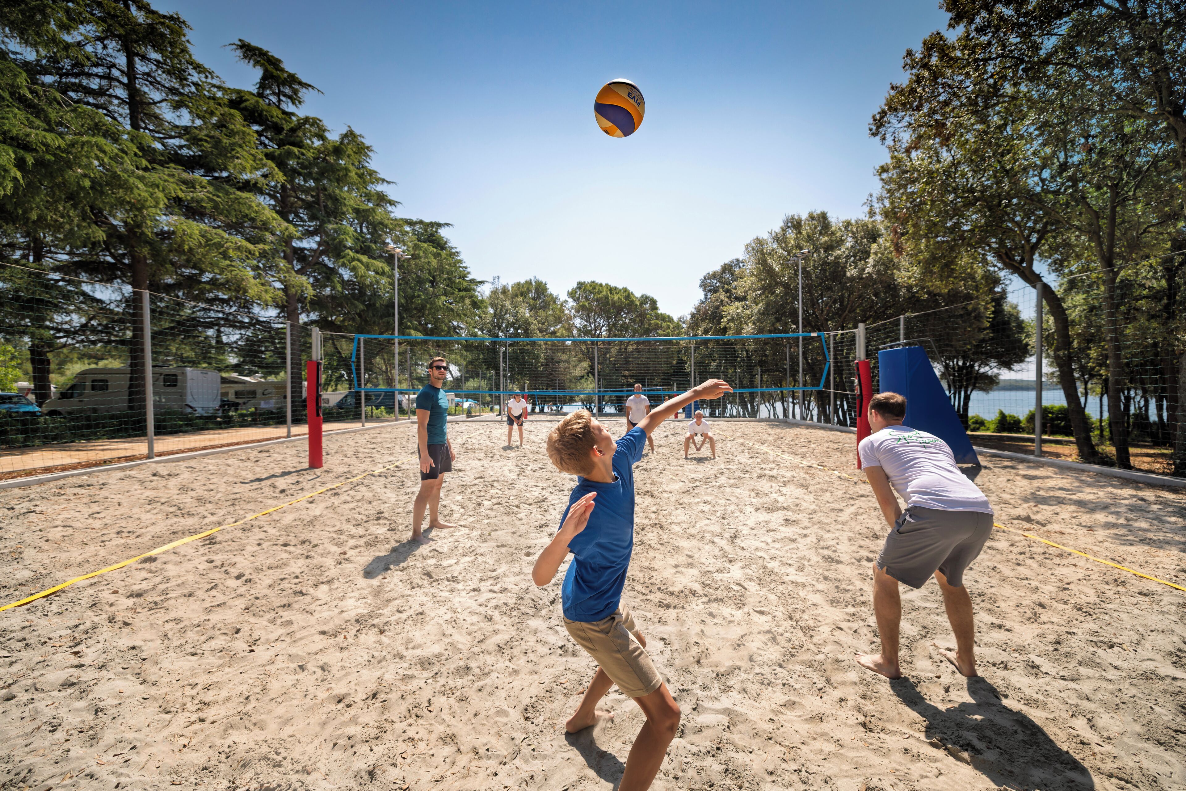 A sandy beach volleyball court with a net, surrounded by trees and a body of water in the background.