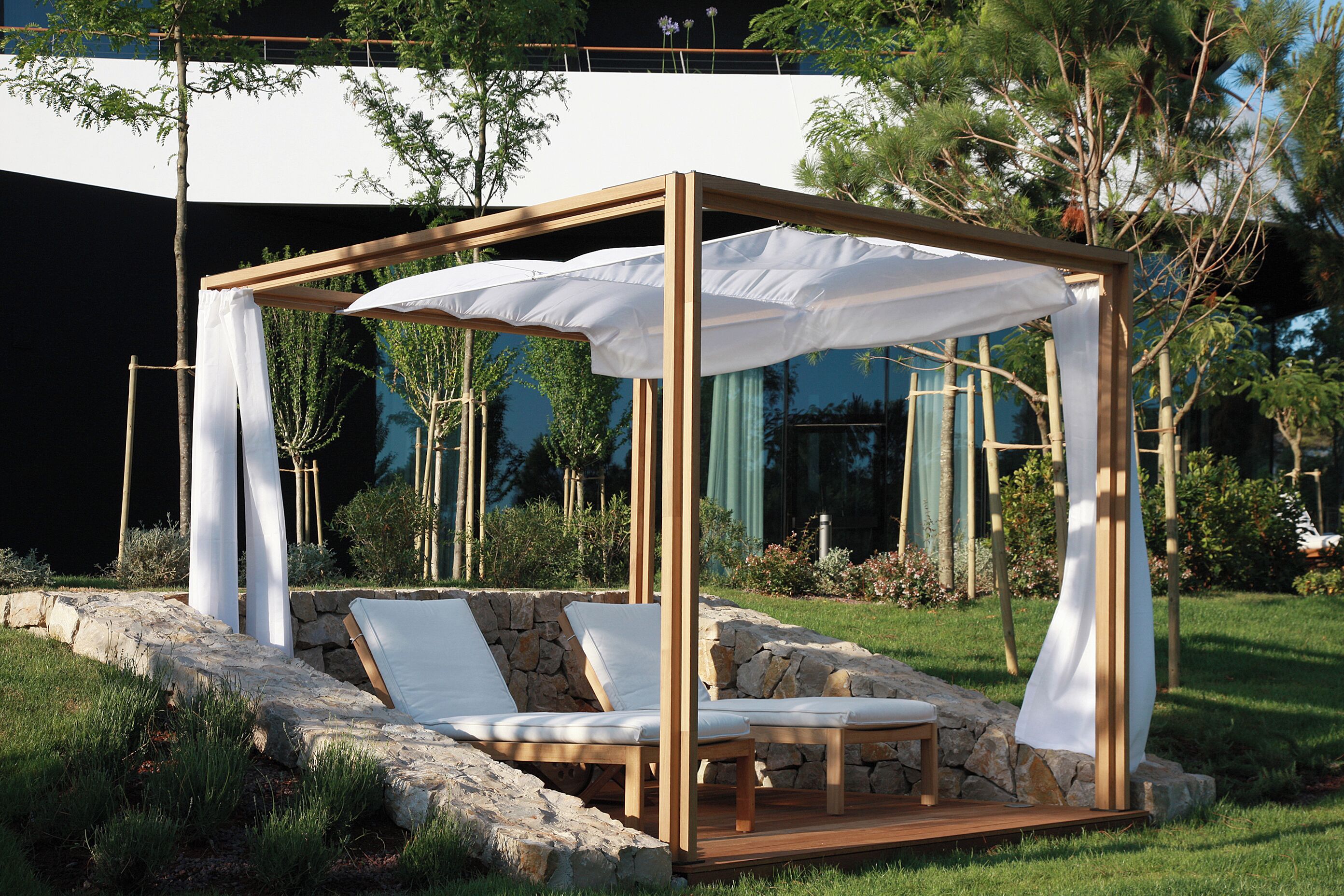 Un gazebo in legno con cuscini bianchi e una tenda, circondato da muri in pietra e vegetazione.