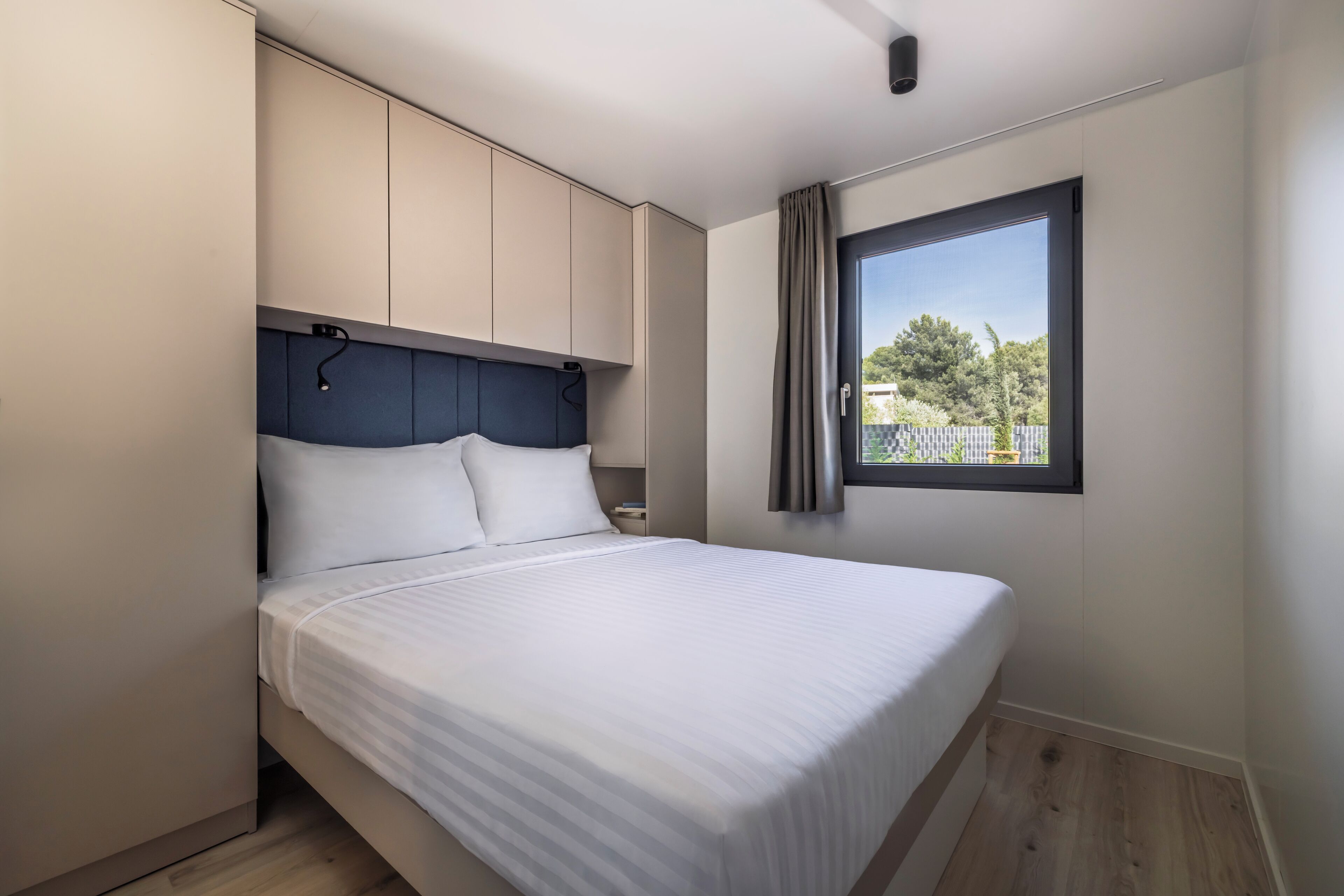 A modern bedroom with a large bed, built-in storage, a window with a view, and a ceiling light.