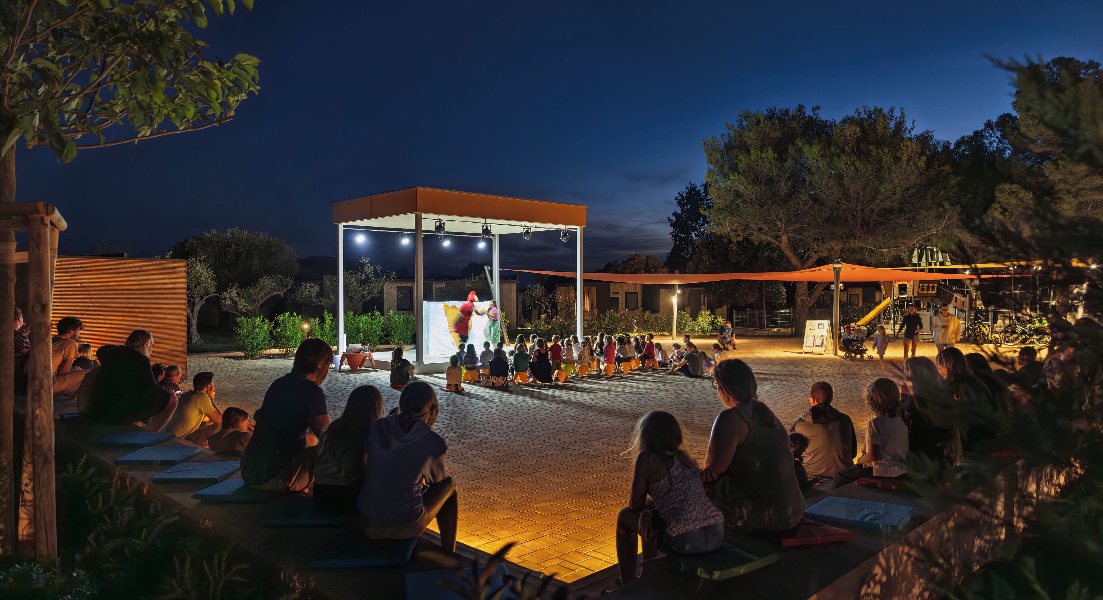 An outdoor gathering area with a covered stage and seating for an audience.