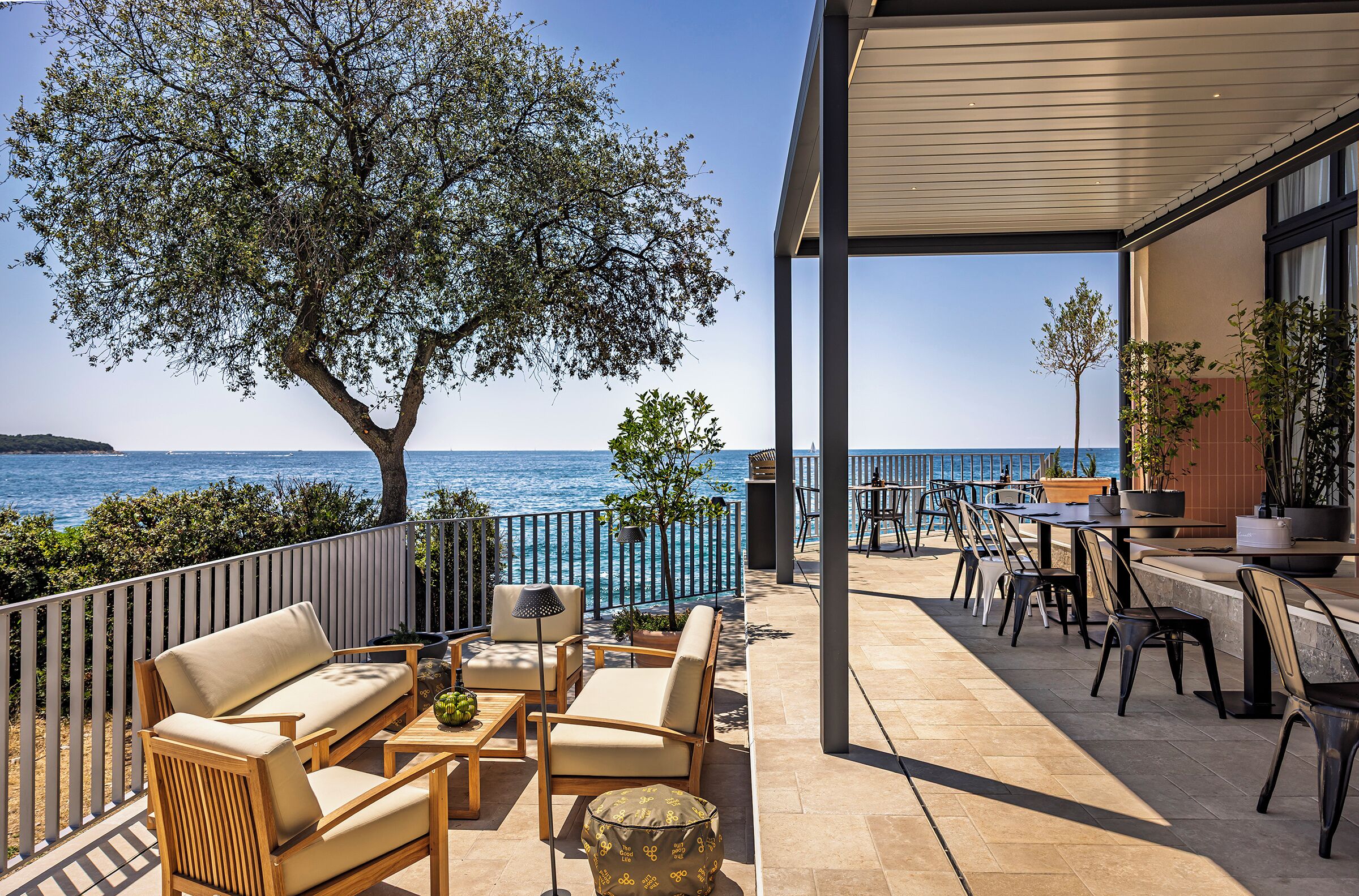A terrace with outdoor seating, a view of the sea, and a tree providing shade.