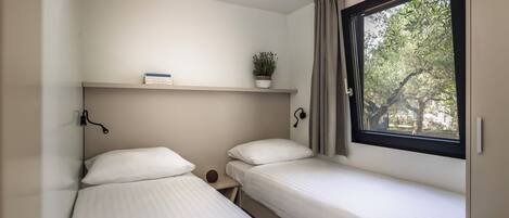 A bedroom with two single beds, a bedside table, a shelf with a plant, and a window with a view of trees.