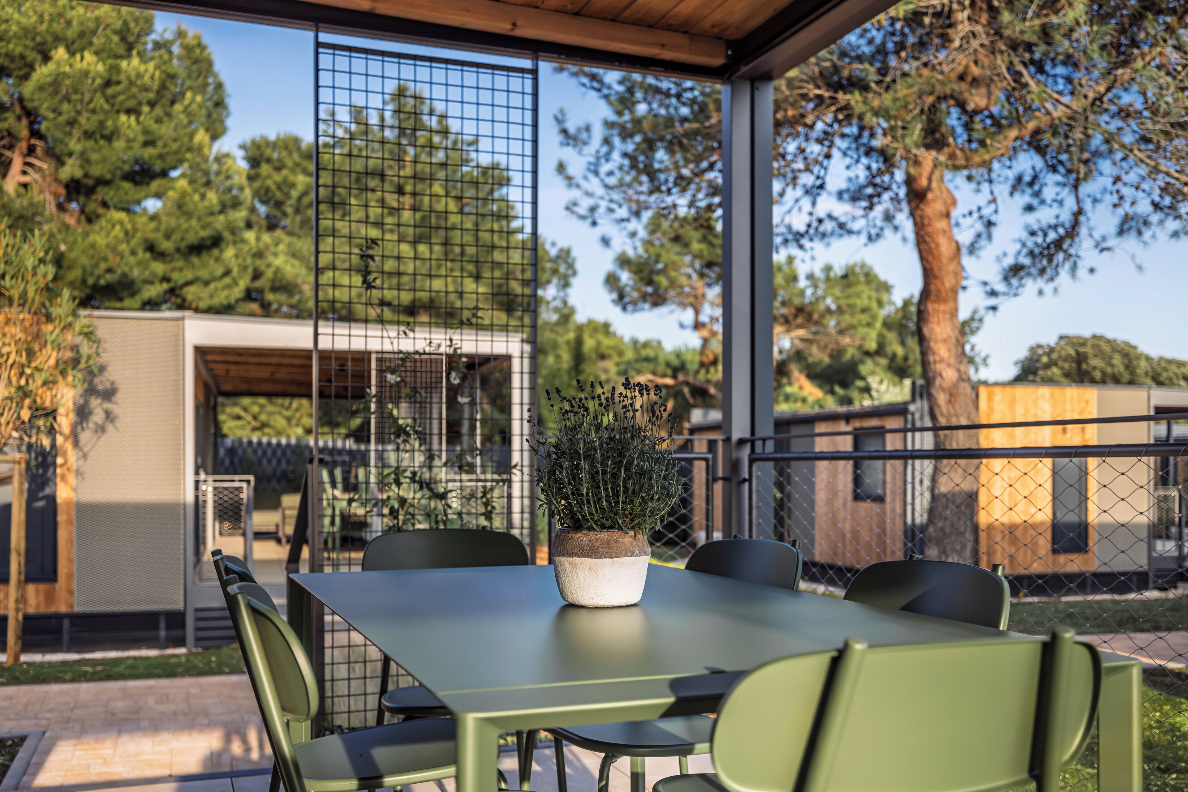 A covered outdoor dining area with a table and chairs, a potted plant, and a view of a building and trees.
