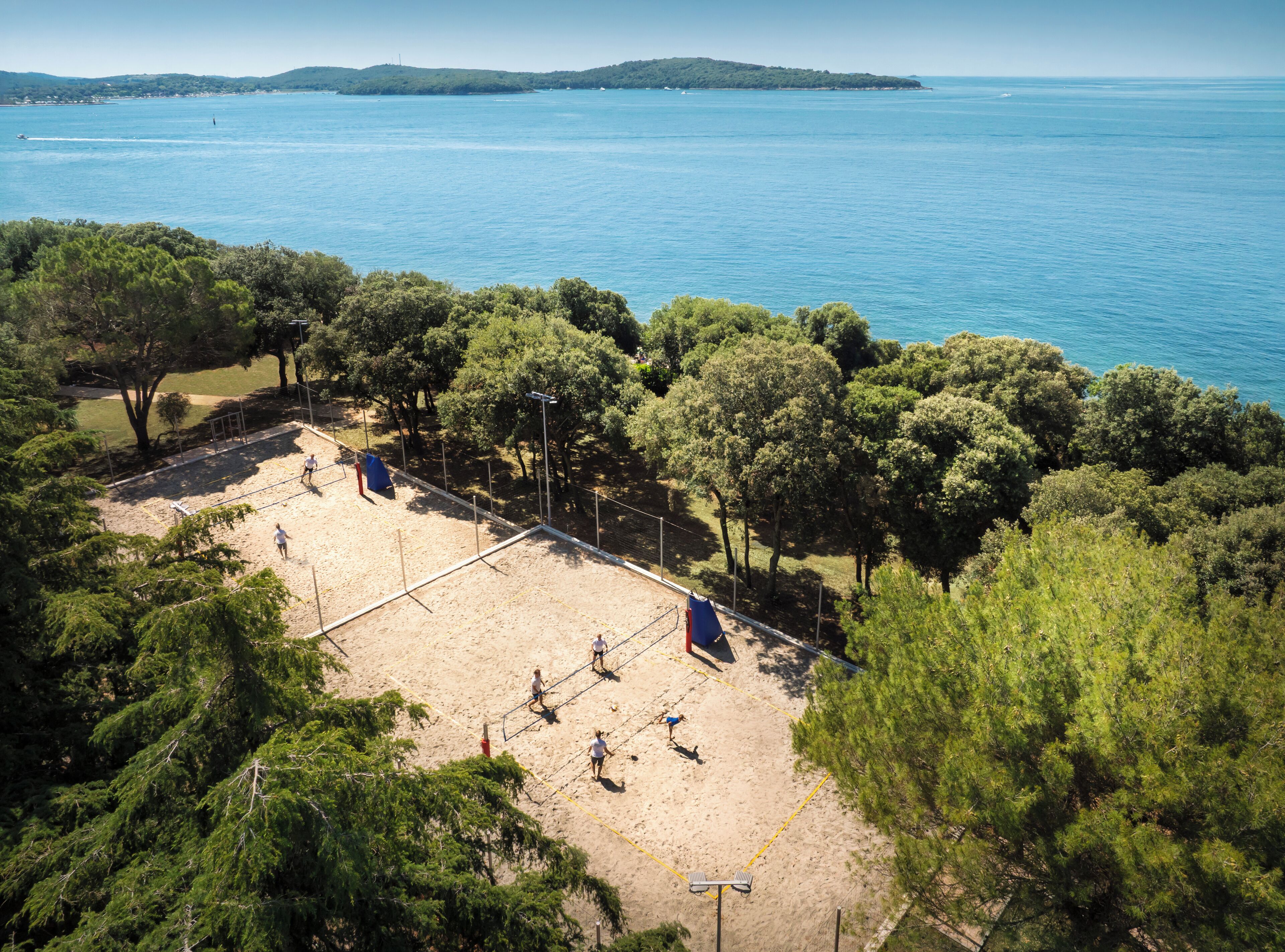 A beach volleyball court surrounded by trees, with a view of the sea and distant hills.