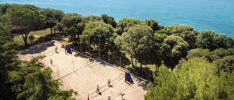 A beach volleyball court surrounded by trees, with a view of the sea and distant hills.