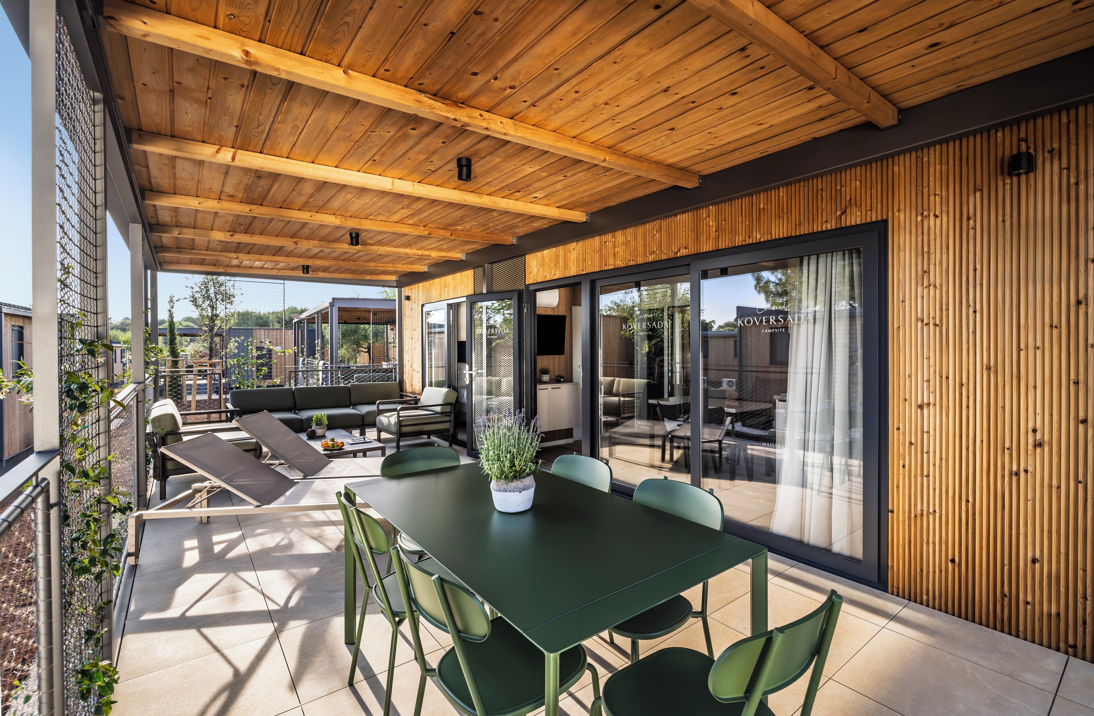 Um pátio externo com teto de madeira, cadeiras verdes, uma mesa e um vaso de planta.