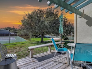 A wooden deck with outdoor furniture, a tennis court, and a tree in the background.