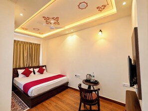 A hotel room with a bed, a small table with a kettle, a chair, and a television mounted on the wall. - KALAKAR HAVELI - Udaipur (Udaipur)