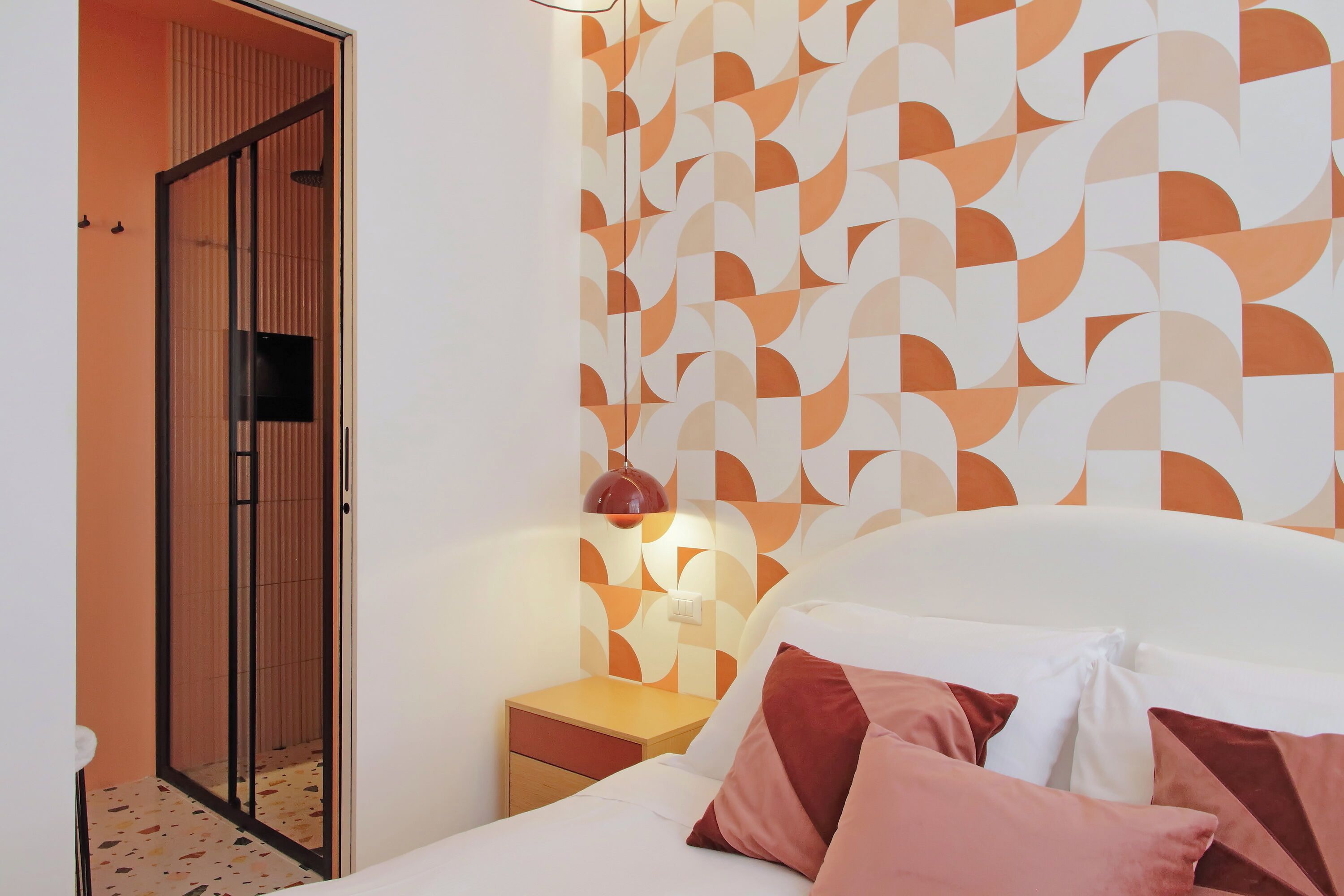 Een slaapkamer met een bed, kussens, een nachtkastje, een lamp en een muur met een geometrisch patroon.
