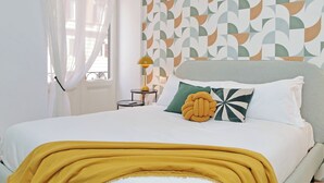 Un lit bien fait, agrémenté d’un plaid couleur moutarde, un mur orné d’un motif décoratif et une petite table de chevet avec une lampe.