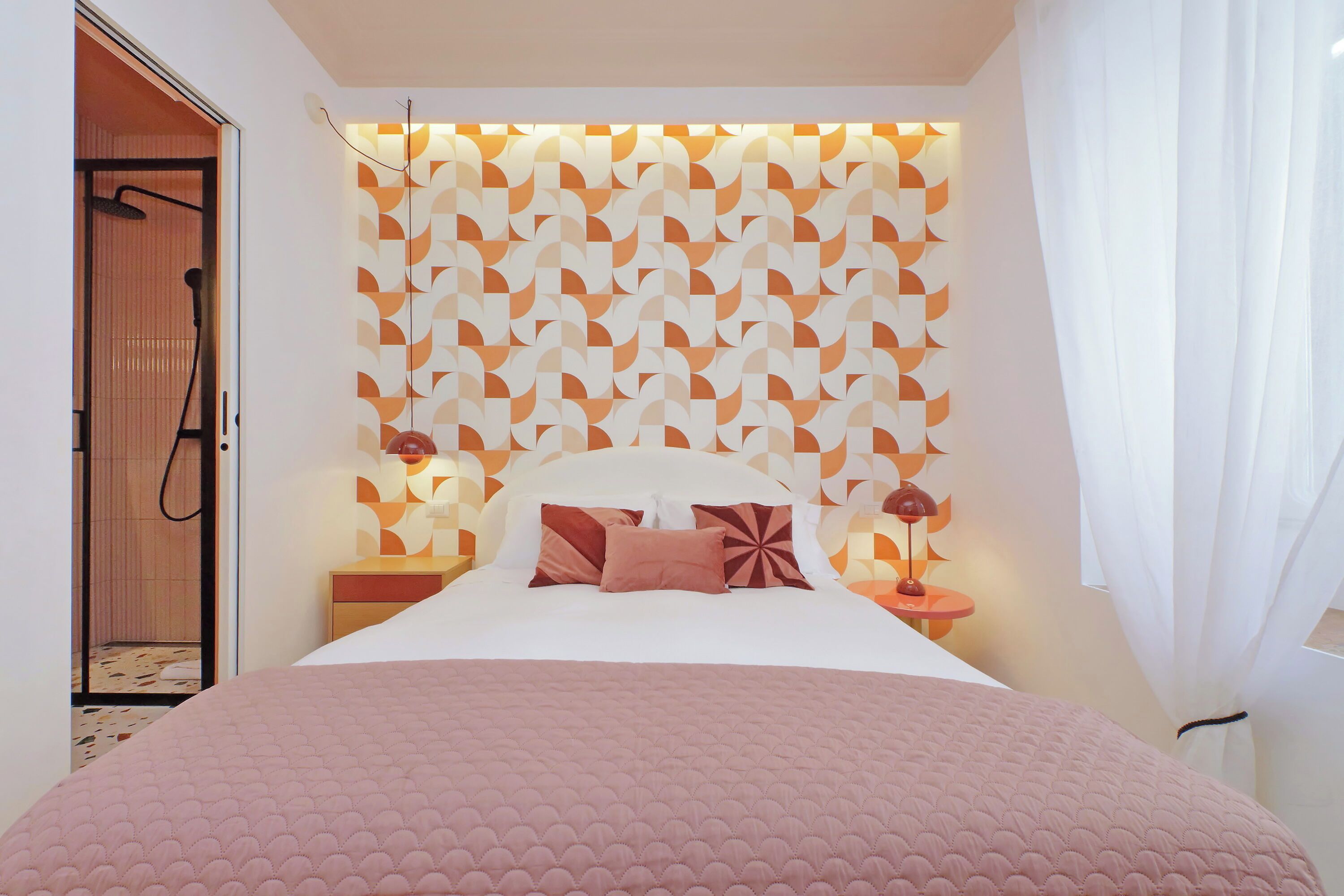 A bedroom with a patterned headboard, a bed with pink bedding, two bedside lamps, and a visible shower area.