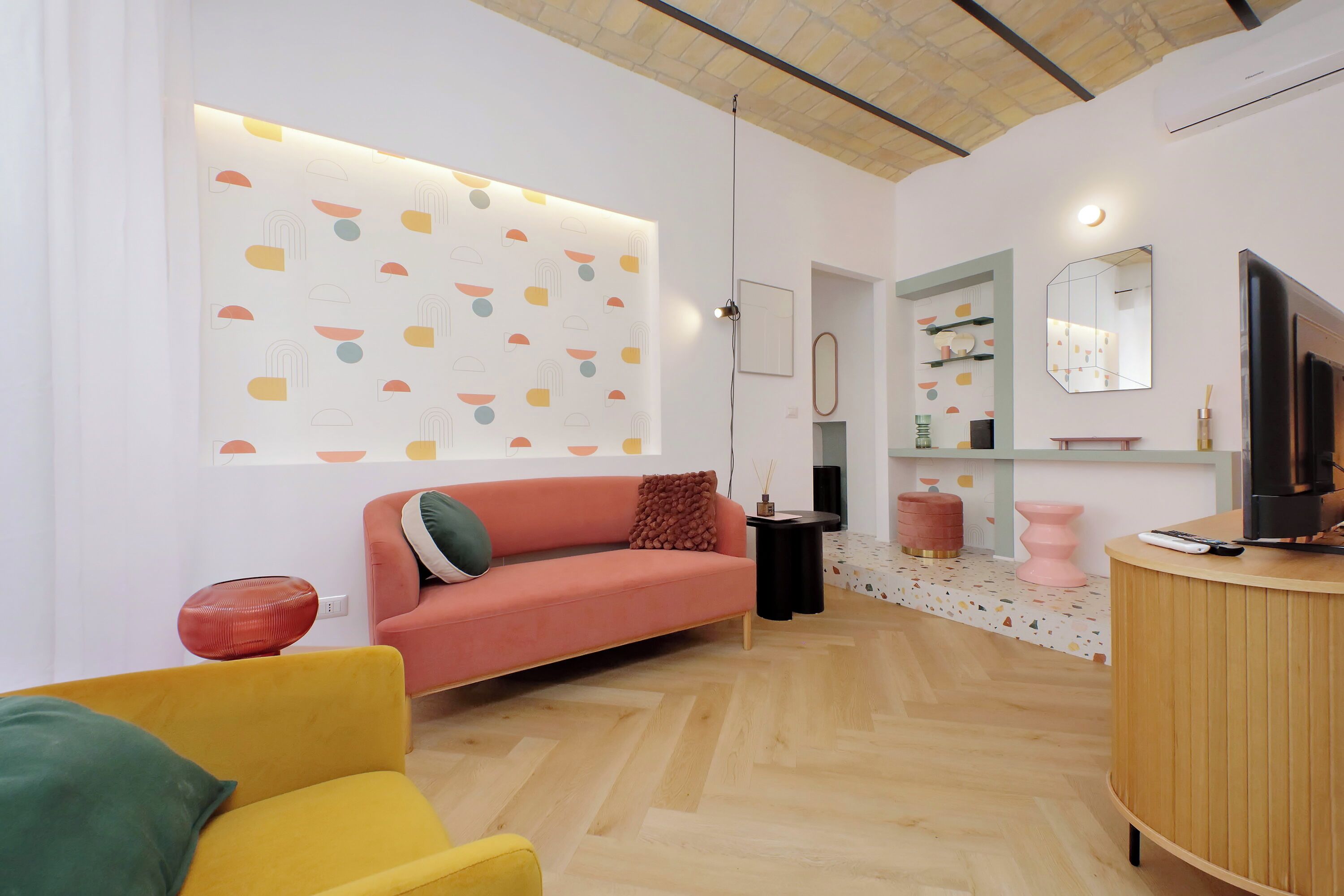 A modern living room with a sofa, a yellow armchair, a wooden cabinet, and a decorative wall with geometric patterns.