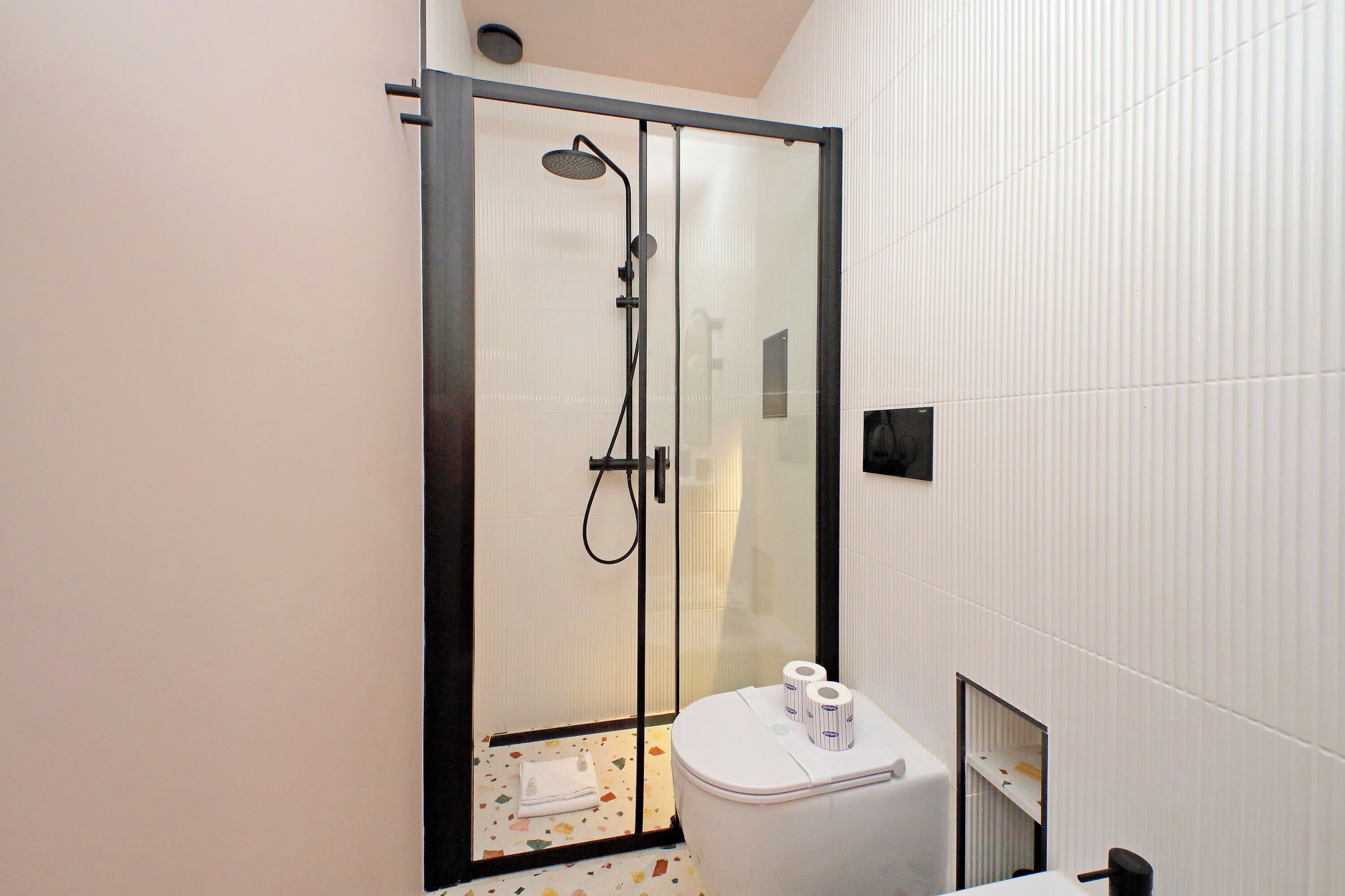 Un baño moderno con ducha de vidrio, un inodoro con papel higiénico y piso de baldosas.