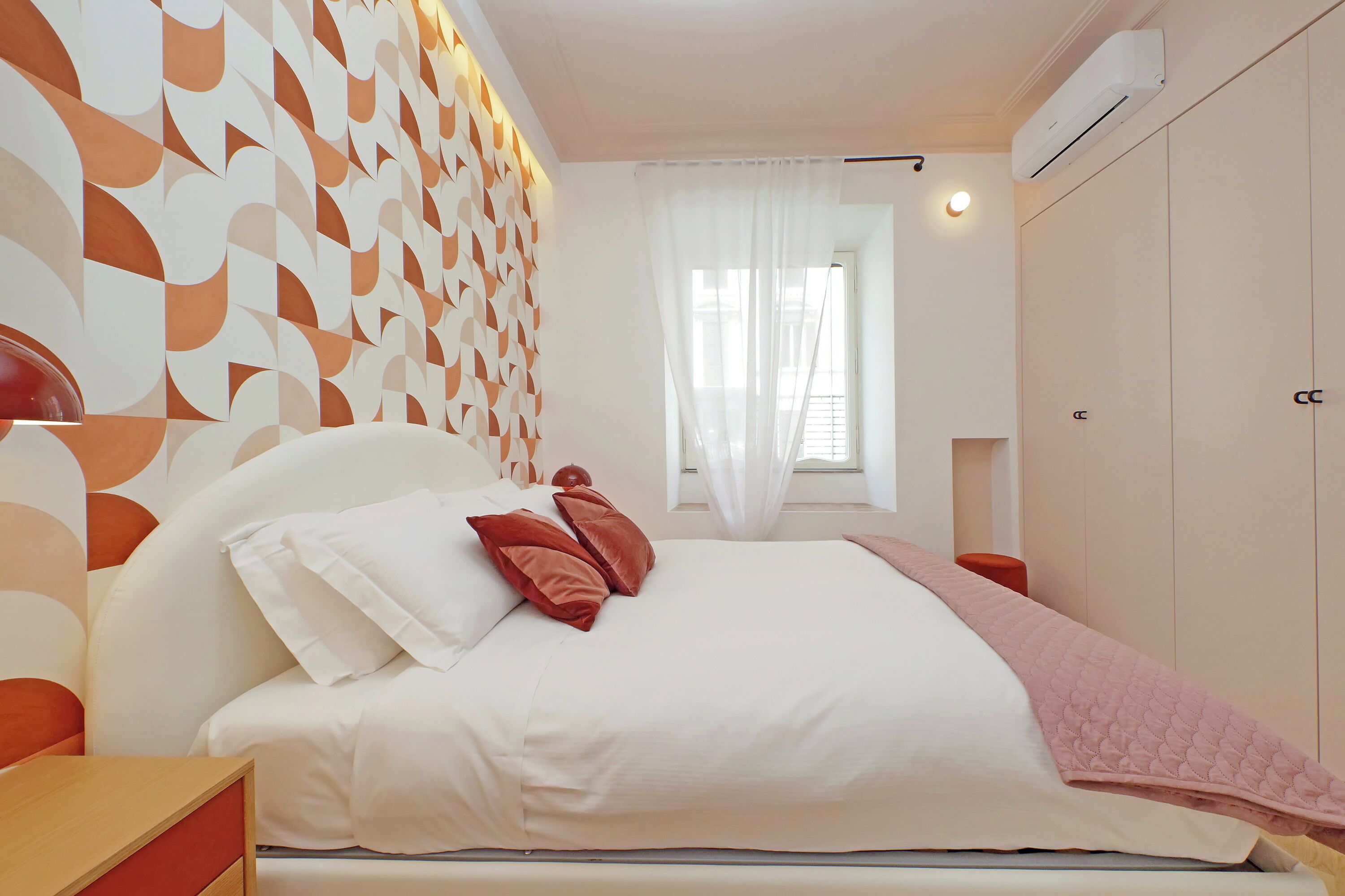 Une chambre à coucher avec un lit, une table de chevet, une fenêtre avec des rideaux et un mur orné d’un motif géométrique.