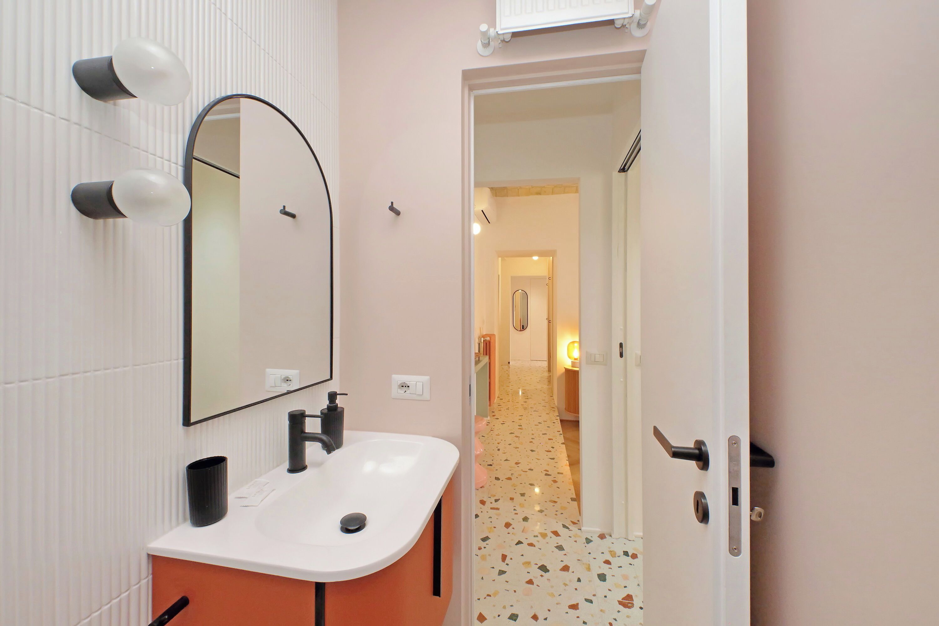 A modern bathroom with a white sink, a large mirror, and a door leading to a hallway with a patterned floor.