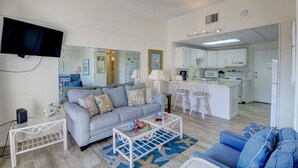A modern living room with a sofa, coffee table, and a TV. There is a kitchen area with white cabinets and a dining space. - 620 El Matador 1 Bedroom Condo (Fort Walton Beach)