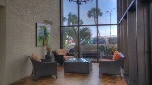 A sunlit indoor space with wicker furniture, a glass coffee table, and a large window offering a view of palm trees and parked cars outside.