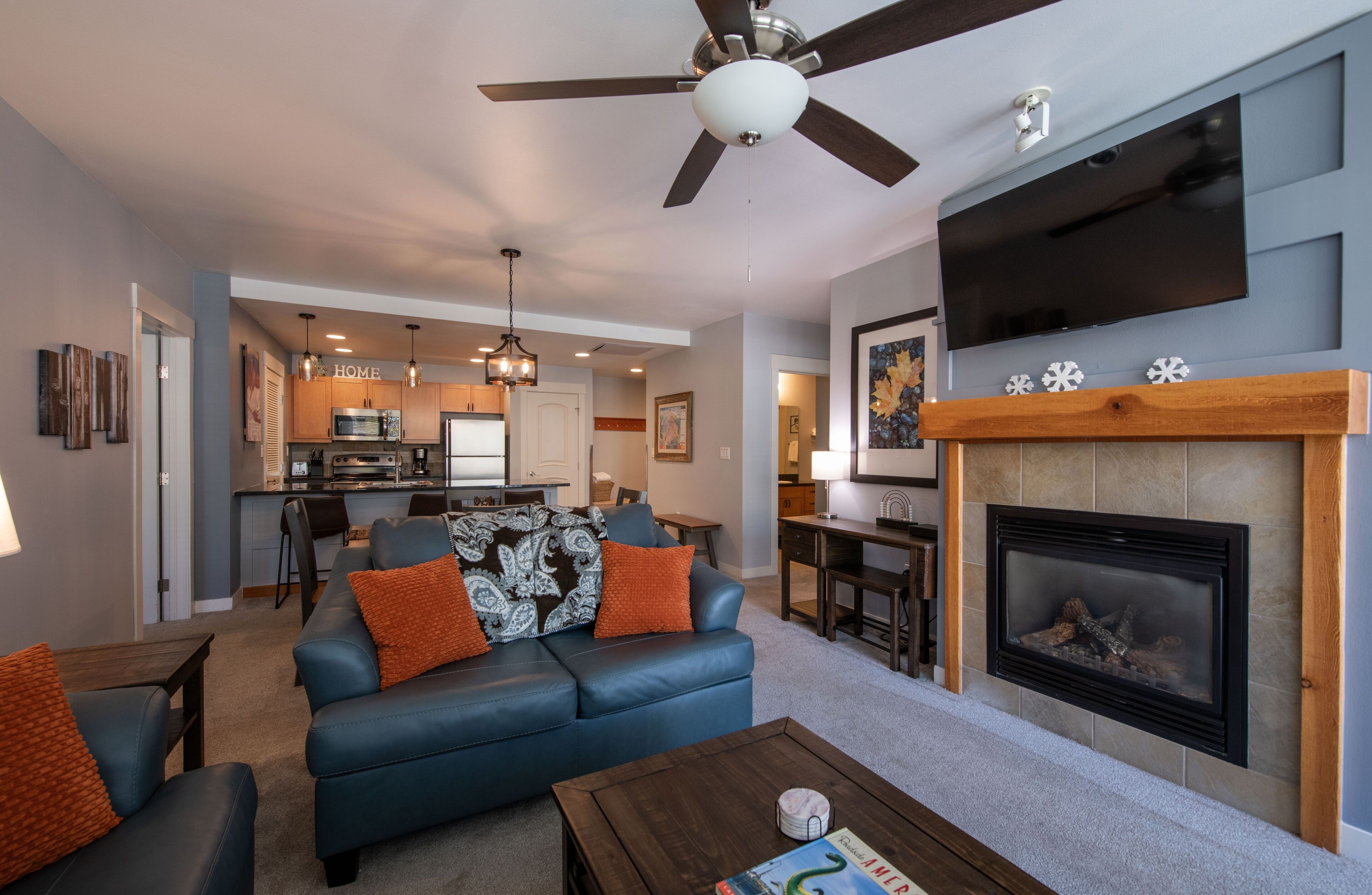 A modern living room with a fireplace, a ceiling fan, and a kitchen in the background.