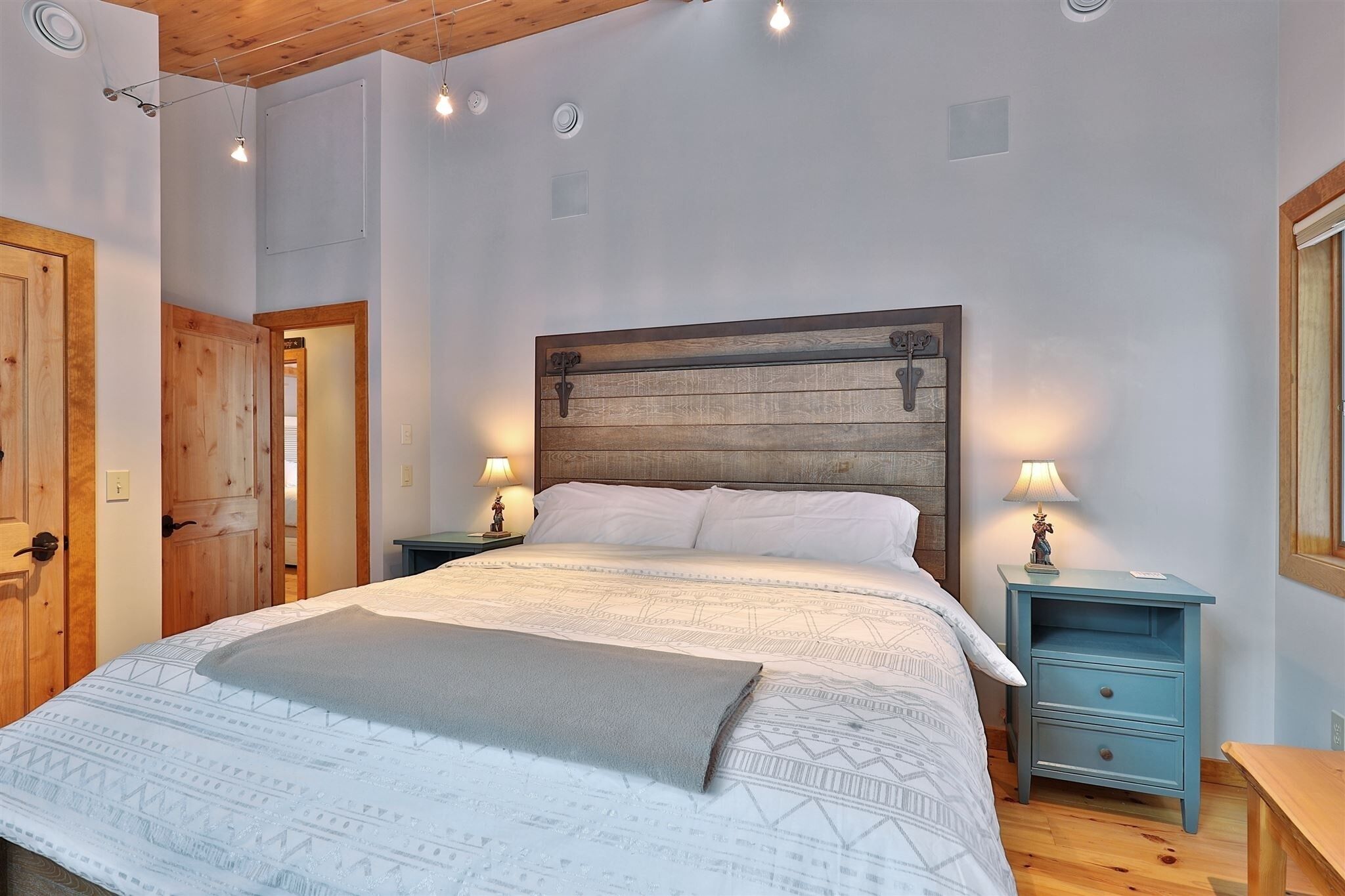 Un dormitorio con cabecera de madera, una cama grande con ropa de cama estampada, dos mesitas de noche con lámparas y puertas de madera.
