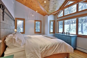 Un dormitorio con techo de madera, ventanas amplias, una cama con ropa blanca y un aparador azul.