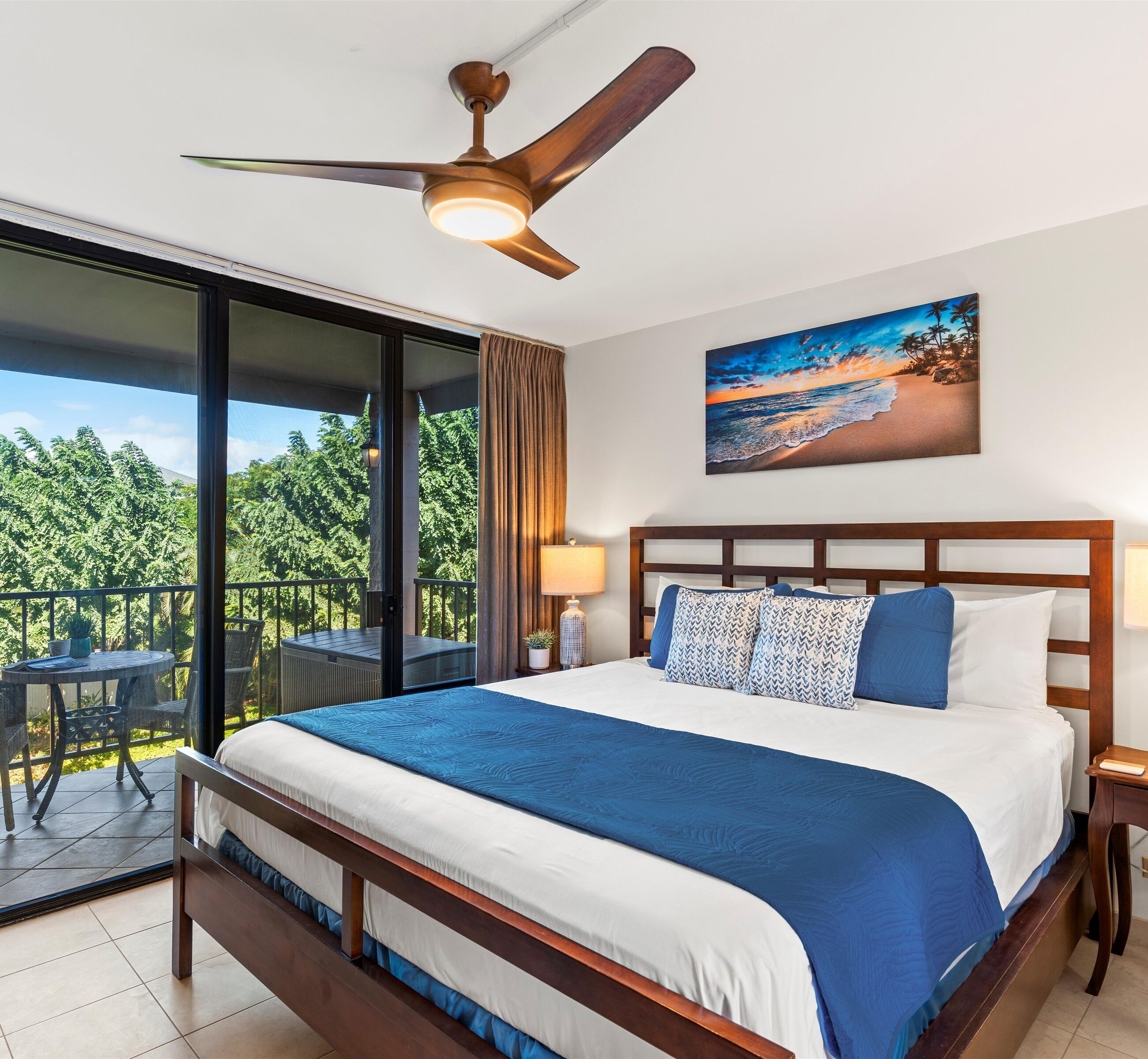 Ein Hotelzimmer mit einem großen Bett, einem Deckenventilator, einem Balkon mit Tisch und Stühlen sowie einem Bild mit einer Strandansicht.