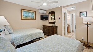 Habitación de hotel con dos camas, ventilador de techo, televisión, aire acondicionado, espejo y un pasillo que conduce a un baño.